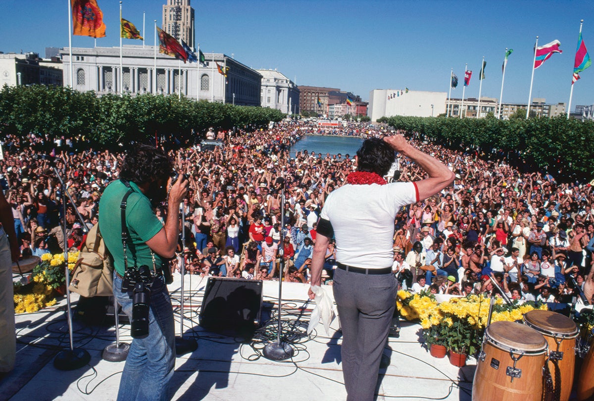 Supervisor Harvey Milk addressing Gay Freedom Day celebrants gathered at United Nations Plaza in San Francisco, with the first rainbow flags visible in the distance  June 25, 1978 