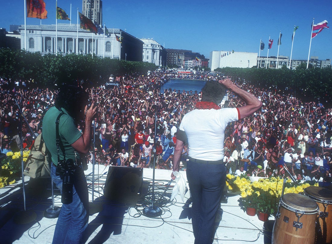 Supervisor Harvey Milk addressing Gay Freedom Day celebrants gathered at United Nations Plaza in San Francisco, with first rainbow flags visible in the distance
