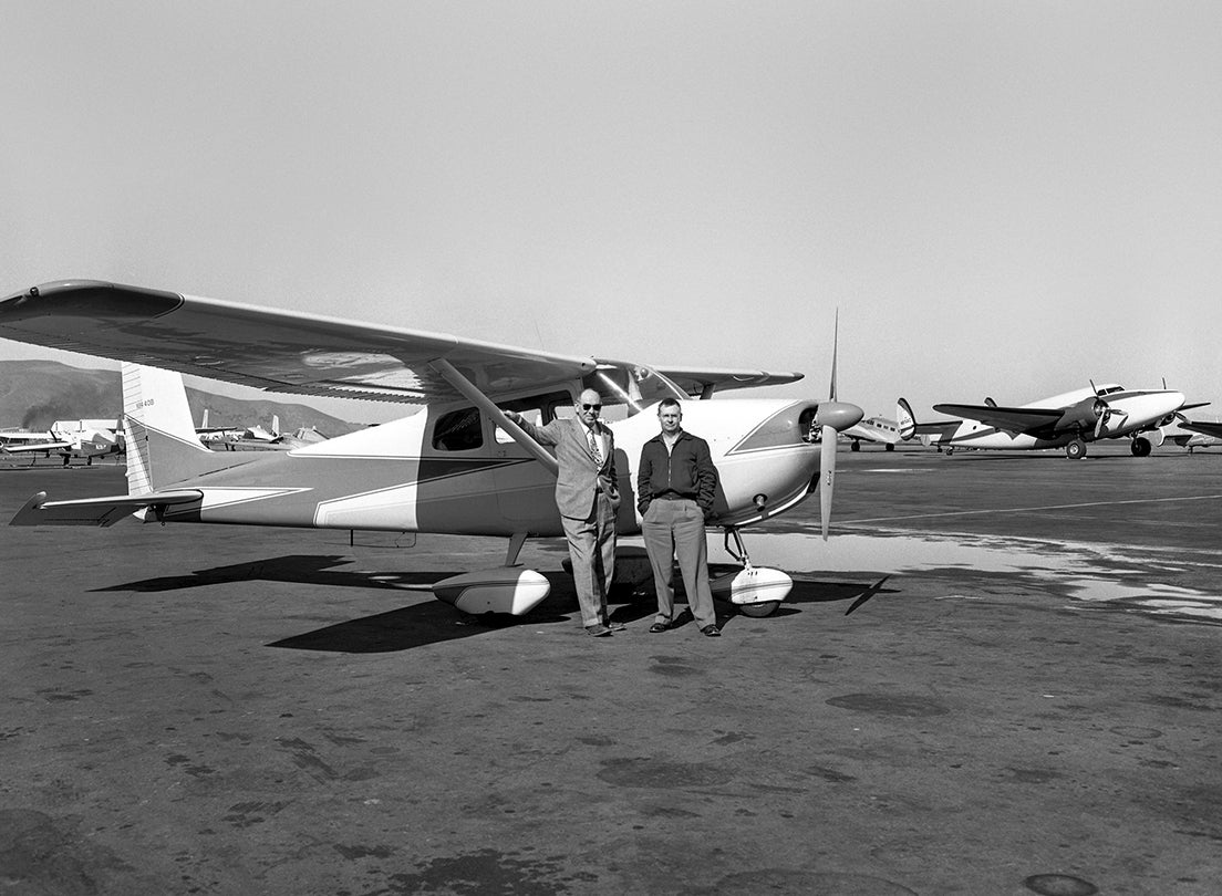 (L-R) Photographers Marshall Moxom and Ken Snodgrass posing in front of a Cessna 175 aircraft, San Francisco International Airport (SFO), February 19, 1960; view facing north with San Bruno Mountain visible on horizon.