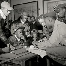 332nd Fighter Group pilots attending a briefing: (front row L-R) unidentified, Jimmie D. Wheeler (with goggles), Emile G. Clifton (cloth cap), (standing L-R) Ronald W. Reeves (cloth cap), Hiram Mann (leather cap), Joseph L. Chineworth (wheel cap) Elwood T. Driver, Edward Thomas (partial view), Woodrow W. Crockett (wheel cap)  March 1945