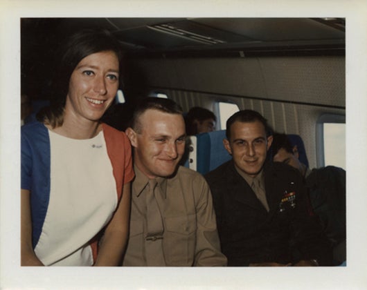 United Air Lines stewardess Sandra Herrmann poses in aircraft cabin with passengers / service members aboard U.S. Military Airlift Command (MAC) charter flight  c. 1968